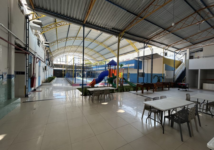 Área de convivência coberta com mesas, playground ao fundo e acesso à quadra